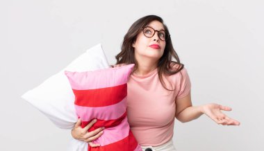 Güzel bir kadın omuz silkiyor, kafası karışık ve kararsız. Pijama giyiyor ve yastık tutuyor.