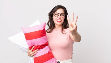 Güzel bir kadın gülümsüyor ve arkadaş canlısı görünüyor. Üç numara pijama giyiyor ve yastık tutuyor.