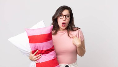 Güzel bir kadın ağzı açık, pijama giymiş ve elinde yastıkla şok olmuş ve şaşırmış görünüyor.