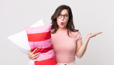 Güzel bir kadın şaşırmış ve şok olmuş görünüyor, çenesi düşmüş pijama giymiş ve elinde yastıkla bir nesne tutuyor.