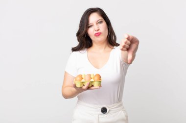 Güzel bir kadın kendini kötü hissediyor, baş parmaklarını gösteriyor ve yumurta kutusunu tutuyor.