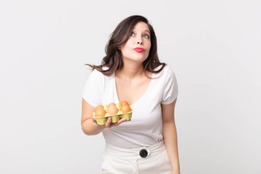 Güzel bir kadın omuz silkiyor, kafası karışık ve kararsız ve elinde bir yumurta kutusu tutuyor.