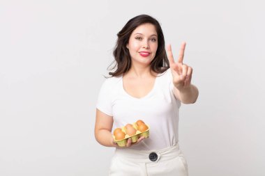 Güzel bir kadın gülümsüyor ve arkadaş canlısı görünüyor. İki numarayı gösteriyor ve elinde bir yumurta kutusu tutuyor.