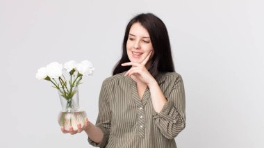 Güzel bir kadın mutlu bir şekilde gülümser ve hayal kurar ya da şüphe eder ve elinde dekoratif bir çiçek taşır. Kulaklıklı bir yardımcı ajan.