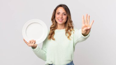 Oldukça zayıf bir kadın gülümsüyor ve arkadaş canlısı görünüyor, dört numarayı gösteriyor ve boş bir tabak tutuyor ve boş bir tabak tutuyor.