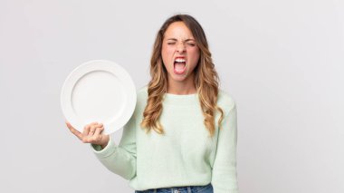 Oldukça zayıf bir kadın agresif bir şekilde bağırıyor, kızgın görünüyor ve elinde boş bir tabakla boş bir tabak tutuyor.