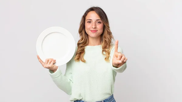 stock image pretty thin woman smiling proudly and confidently making number one and holding an empty plate and holding an empty dish