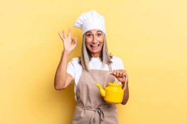 Orta yaşlı aşçı kadın kendini mutlu, rahat ve tatmin olmuş hissediyor. Onayını iyi bir jestle gösteriyor. Gülümsüyor ve elinde çaydanlık tutuyor.