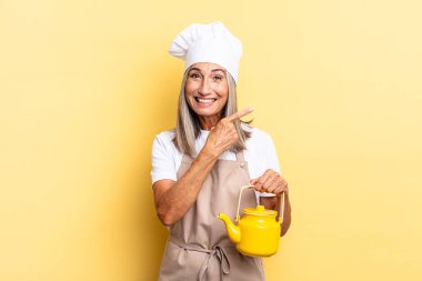 Orta yaşlı aşçı kadın heyecanlı ve şaşırmış görünüyor. Kenarlarını ve yukarıyı göstererek yeri kopyalıyor ve elinde bir çaydanlık tutuyor.