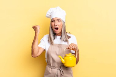 Orta yaşlı aşçı kadın sinirli bir ifadeyle bağırıyor ya da yumruklarını sıkıp başarıyı kutluyor ve elinde çaydanlık tutuyor.