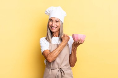Orta yaşlı aşçı kadın mutlu, pozitif ve başarılı hissediyor, meydan okumayla karşılaştığında motive oluyor veya iyi sonuçları kutluyor ve elinde boş bir demlik tutuyor.
