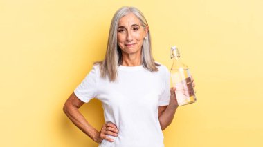 Mutlu bir şekilde gülümsüyor, eli kalçasında ve kendinden emin, pozitif, gururlu ve arkadaş canlısı bir tavır içinde ve elinde bir su şişesi tutuyor.