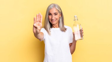 Gülümsüyor ve arkadaş canlısı görünüyor, 4 ya da 4 numarayı gösteriyor, geri sayıyor ve su şişesini tutuyor.