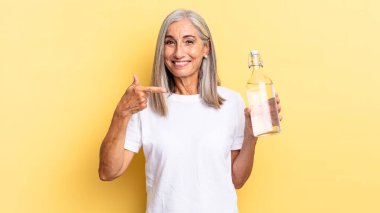 Neşeli bir şekilde gülümseyen, mutlu hisseden, kenarı ve yukarıyı gösteren, fotokopi uzayındaki nesneleri gösteren ve su şişesini tutan