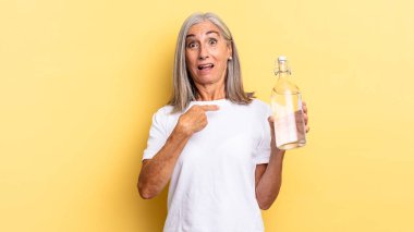 Şok olmuş ve şaşırmış bir halde, ağzı açık, kendini işaret eden ve su şişesini tutan.