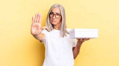 Ciddi görünüyor, sert, hoşnutsuz ve kızgın. Avuç içini açıp jesti durduruyor ve elinde beyaz bir kutu tutuyor.