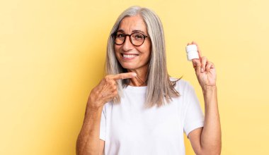 Neşeli bir şekilde gülümsüyor, mutlu hissediyor ve yan ve yukarı doğru işaret ediyor, fotokopi alanındaki nesneleri gösteriyor ve elinde bir hap şişesi tutuyor.