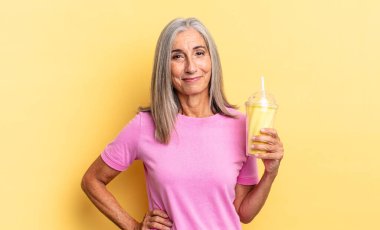 Mutlu bir şekilde gülümsüyor, eli kalçasında ve kendinden emin, pozitif, gururlu ve arkadaş canlısı bir tavır içinde ve elinde bir milkshake tutuyor.