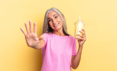 Gülümsüyor ve arkadaş canlısı görünüyor, 5 ya da 5 numarayı gösteriyor, geri sayıyor ve milkshake tutuyor.