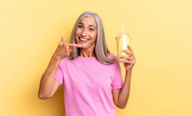 Heyecanlı ve şaşırmış görünüyor. Kenarları ve yukarıyı göstererek boşluğu kopyalıyor ve elinde milkshake tutuyor.