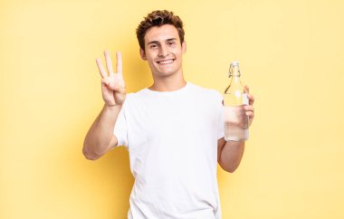 Gülümsüyor ve dost canlısı görünüyor. Üç ya da üç numarayı gösteriyor. Elleri önde, geriye doğru sayıyor. su şişesi kavramı