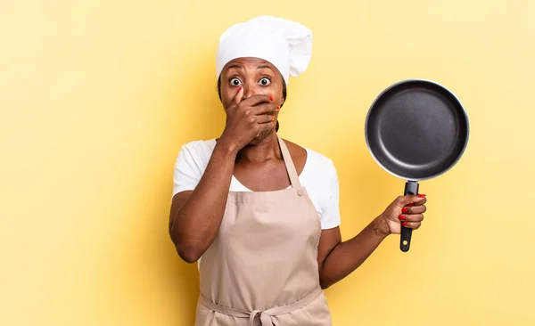 black afro chef woman covering mouth with hands with a shocked, surprised expression, keeping a secret or saying oops
