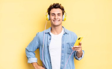 Mutlu bir şekilde gülümsüyor, eli kalçasında ve kendinden emin, olumlu, gururlu ve arkadaş canlısı bir tavırla. kulaklıklar ve akıllı telefon konsepti