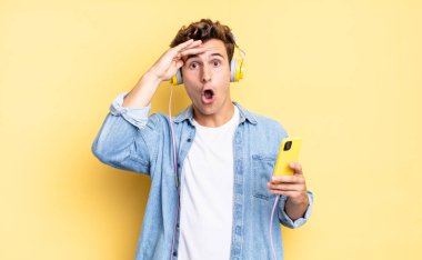 Mutlu, şaşkın ve şaşırmış görünüyor, gülümsüyor ve inanılmaz ve inanılmaz iyi haberleri fark ediyor. kulaklıklar ve akıllı telefon konsepti