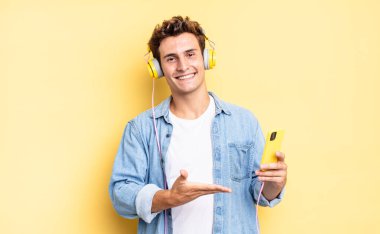 Neşeli bir şekilde gülümseyen, mutlu hisseden ve fotokopi uzayında avucunun içinde bir konsept gösteren. kulaklıklar ve akıllı telefon konsepti