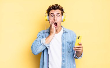 Şok olmuş ve korkmuş hissediyorum, ağzı açık ve elleri yanaklarında korkmuş görünüyor. kulaklıklar ve akıllı telefon konsepti