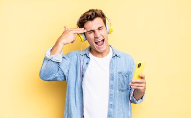 Mutsuz ve stresli görünüyor, intihar jesti elle silah işareti yapıyor, kafasını işaret ediyor. kulaklıklar ve akıllı telefon konsepti