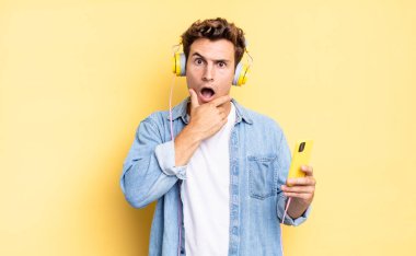 Ağzı ve gözleri sonuna kadar açık ve elleri çenesinde, hoş olmayan bir şokta, ne ya da vay canına diyor. kulaklıklar ve akıllı telefon konsepti