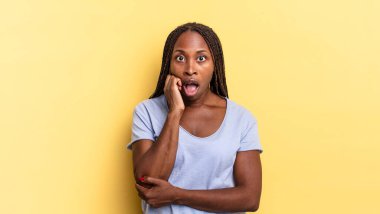 Afro siyah güzel bir kadın şok ve inançsızlıkla ağzı açık, elleri yanağında ve kolları çapraz, sersemlemiş ve şaşırmış hissediyor.