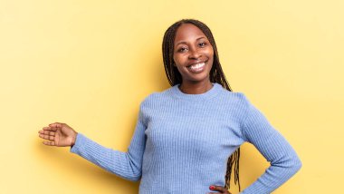 Afro siyah güzel kadın mutlu ve neşeli hissediyor, gülümsüyor ve hoş karşılıyor, dostça bir jestle seni içeri davet ediyor.