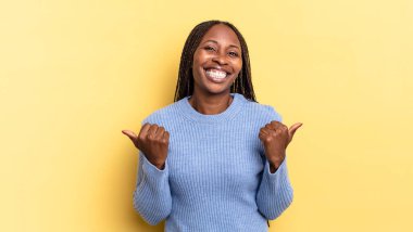 Afro siyah güzel bir kadın neşeyle gülümsüyor ve mutlu görünüyor, kaygısız ve pozitif hissediyor iki başparmağıyla