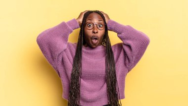 Afro siyah güzel bir kadın heyecanlı ve şaşırmış görünüyor, ağzı açık, iki eli başında, kendini şanslı bir kazanan gibi hissediyor.