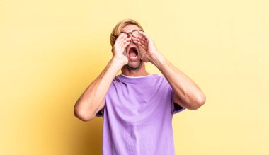 Yakışıklı, sarışın, yetişkin bir adam mutlu, heyecanlı ve pozitif hissediyor. Elleri ağızda, bağırıyor.