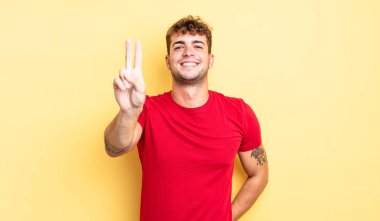Genç yakışıklı adam gülümsüyor ve mutlu görünüyor, kaygısız ve olumlu, zafer veya barış işareti yapıyor.