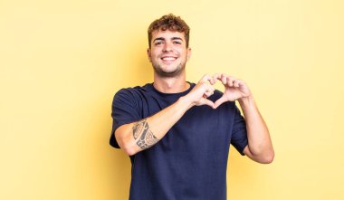 Genç yakışıklı adam gülümsüyor ve mutlu, sevimli, romantik ve aşık, iki eliyle kalbi şekillendiriyor.