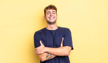 Genç yakışıklı adam kollarını kavuşturup mutlu bir şekilde gülüyor, rahat, pozitif ve tatmin edici bir pozla.