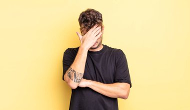 Genç ve yakışıklı bir adam stresli, utanmış ya da üzgün görünüyor, baş ağrısı var, yüzünü eliyle kapatıyor.