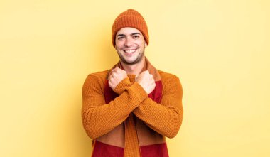 Genç yakışıklı adam, neşeli bir şekilde gülümsüyor ve kutluyor. Yumruklarını sıkmış, kollarını kavuşturmuş, mutlu ve pozitif hissediyor.