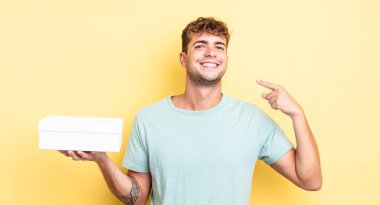 Genç ve yakışıklı bir adam kendi gülüşünü işaret ederek kendine güvenerek gülümsüyor. beyaz kutu kavramı