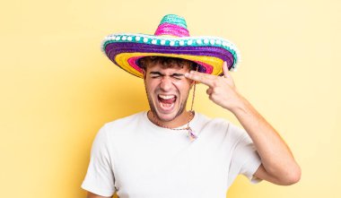 Genç yakışıklı adam mutsuz ve stresli görünüyor, intihar jesti silah işareti yapıyor. Meksika şapkası konsepti