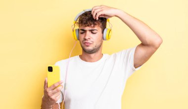 Genç yakışıklı adam mutlu bir şekilde gülümsüyor ve hayal kuruyor ya da şüphe ediyor. kulaklıklar ve akıllı telefon konsepti
