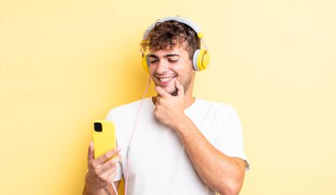 Genç ve yakışıklı bir adam mutlu ve kendinden emin bir ifadeyle gülümsüyor, elleri çenesinde. kulaklıklar ve akıllı telefon konsepti
