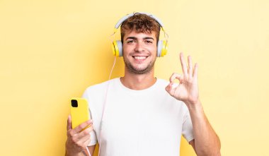 Genç yakışıklı adam kendini mutlu hissediyor, iyi bir jestle onaylıyor. kulaklıklar ve akıllı telefon konsepti
