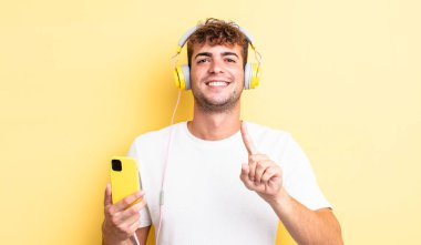 Genç yakışıklı adam gururla gülümsüyor ve kendinden emin bir numara oluyor. kulaklıklar ve akıllı telefon konsepti