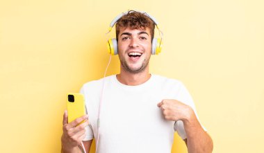 Genç ve yakışıklı bir adam mutlu hissediyor ve heyecanla kendini işaret ediyor. kulaklıklar ve akıllı telefon konsepti