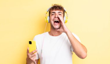 Genç yakışıklı adam mutlu hissediyor, elleri ağzının kenarında bağırıyor. kulaklıklar ve akıllı telefon konsepti